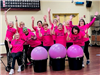 Women in pink shirts with their hands up in the air with three big buckets that have pink exercise balls in them.